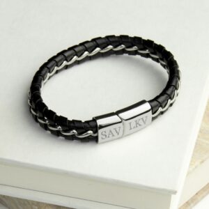 A close-up studio shot of a men’s black leather bracelet featuring a unique silver metal chain interwoven through the braided leather. The bracelet is shown resting on a stack of white books. The polished stainless steel magnetic clasp is the focal point, engraved with the initials "SAV" and "LKV" in a classic serif font.
