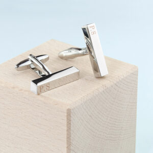 Pair of polished silver bar-shaped cuffhttps://www.google.com/search?q=links with a modern architectural design, featuring custom engraved initials P.S, displayed on a natural light wood block.