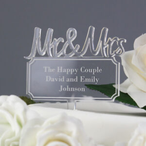 Wide shot of a modern, clear acrylic wedding cake topper, featuring the text "Mr & Mrs" in a flowing script font. It is personalised with an engraved plaque below reading "The Happy Couple David and Emily Johnson." It is set within white roses on a white cake.