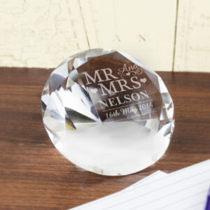 A large, brilliant-cut clear glass diamond paperweight resting on a dark wooden desk. The flat top surface is engraved with "MR AND MRS NELSON" and the date "16th May 2014" in an elegant white font. In the background, vintage-style maps and white lined paper are visible.