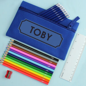 A royal blue microfibre pencil case featuring the name "TOBY" in a bold black frame. A variety of multi-coloured crayons and navy blue pencils, each individually personalised with the name "TOBY", are arranged neatly on a light blue background next to a white 6-inch ruler and a red sharpener.