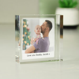 A freestanding square acrylic photo block featuring a high-quality print of a father kissing his baby's forehead. The bottom of the frame includes personalised text reading "Love you Daddy, Jacob x" in a clean black font. The block is set on a white surface with a soft-focus background of a plant.