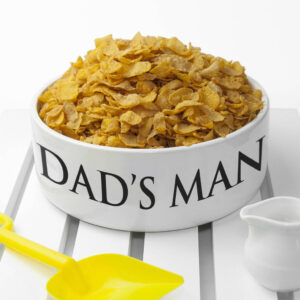 A super large white ceramic bowl overflowing with cornflakes, featuring bold black personalised text that reads "DAD'S MAN BOWL" sitting on a white slatted surface next to a yellow toy spade and a small white milk jug.