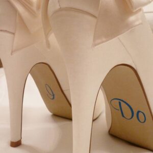 Close-up of ivory high-heeled wedding shoes with silver vinyl stickers on the arches reading "I Do"