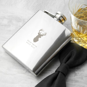 Polished stainless steel 6oz hip flask featuring an engraved stag head motif and custom text, styled on a grey surface next to a glass of whisky and a black bow tie.