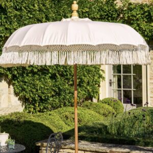 A large cream-coloured canvas teak parasol with long, intricate crochet cotton tassels hanging from the edges. The parasol features a natural wood pole and is standing in a lush, green English garden setting next to a stone wall and manicured hedges.