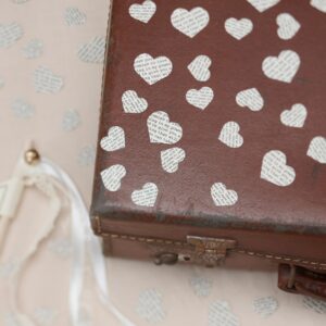 Vintage heart-shaped confetti with printed love poem text scattered over a rustic brown leather suitcase with white ribbons.