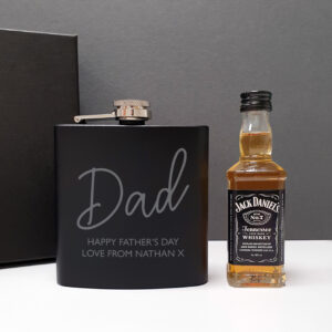 Personalised Hip Flask Gift Set. A sleek matte black 6oz hip flask engraved with the name "Dad" in script and the message "HAPPY FATHER’S DAY LOVE FROM NATHAN X," standing next to a 5cl miniature bottle of Jack Daniel's whisky and a black presentation gift box.