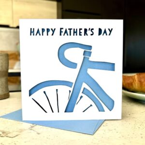 Cycling Father's Day Card: A square Father’s Day card featuring a white laser-cut silhouette of the front half of a road bicycle against a light blue background. The card reads "HAPPY FATHER'S DAY" in stencil-style lettering and is held over a matching blue envelope next to a coffee cup and croissant.