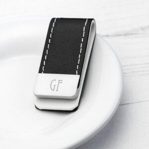 A close-up studio shot of a sleek black PU leather money clip with cream stitching, resting on a white plate. The stainless steel clip section is engraved with the initials "GF" in a Modern Tall font.