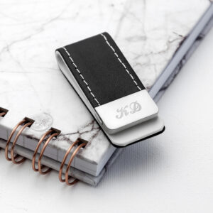 A black leather money clip with cream stitching resting on a notebook. The stainless steel clip is engraved with the initials "KD" in an elegant Script font.