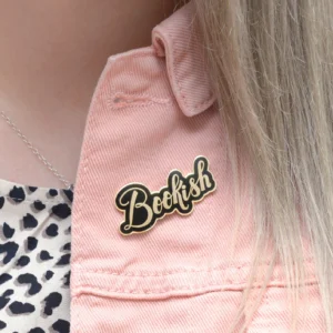 A close-up photograph of a person wearing a pink denim jacket, featuring a shiny black and gold hard enamel lapel pin that reads "Bookish" in elegant cursive script.