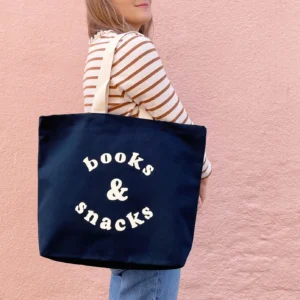 A person wearing a striped shirt carrying a navy blue canvas tote bag that reads "books & snacks" in bold off-white lettering, set against a pink wall background.