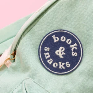 A close-up photograph of a light, mint green textured canvas bag featuring a circular navy blue embroidered patch. The patch contains white text that reads "books & snacks" in a fun, rounded font with an ampersand in the center. A brass-colored zipper pull is partially visible on the left side of the frame against the fabric.