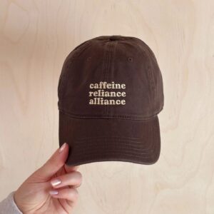 A person holding a dark brown, unstructured cotton baseball cap with the words Caffeine Reliance Alliance embroidered in cream thread on the front.