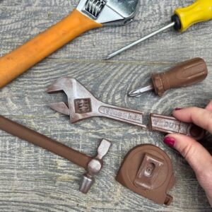 Realistic Chocolate Tool Kit: A set of milk chocolate tools including an adjustable spanner, hammer, and screwdriver. The chocolate tools feature a silver lustre finish and are shown alongside real metal tools on a wooden background to show their authentic detail.