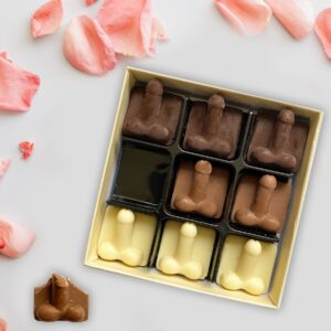Novelty Chocolate Willies: A gold gift box containing nine Belgian chocolates shaped like penises in dark, milk, and white chocolate, arranged on a grey surface with pink rose petals scattered around. One milk chocolate piece is shown broken at the bottom.