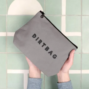 A pair of hands holding a grey canvas wash bag with the word "DIRTBAG" printed in black, set against a light green tiled background.