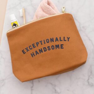 A flat-lay shot of a tan canvas wash bag with 'Exceptionally Handsome' printed in navy blue, sitting on a marble surface with a toothbrush, toothpaste, and a pink face cloth peeking out from the gold zip opening.