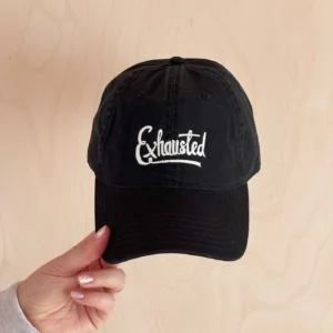 A black cotton baseball cap held in a hand, featuring the word "Exhausted" embroidered in white thread against a neutral wooden background.