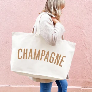 A person wearing a cream coat carrying an oversized natural canvas tote bag with the word Champagne printed in metallic bronze on the front, walking in front of a pink wall.