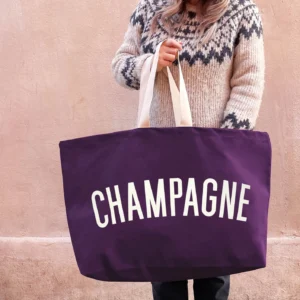 An oversized plum-coloured canvas tote bag with the word Champagne printed in cream on the front, held by someone wearing a patterned jumper.