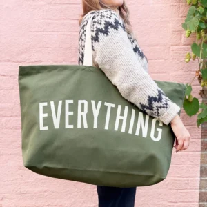 A person wearing a patterned sweater carrying an extra-large khaki green canvas tote bag that reads "EVERYTHING" in bold off-white lettering, set against a pink wall background.