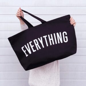 A person standing against a white wooden paneled wall holding a large black canvas tote bag with the word 'EVERYTHING' printed in big white letters.