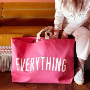 A person sitting on a sofa holding an extra large bright pink canvas tote bag with the word 'EVERYTHING' printed in large white letters.
