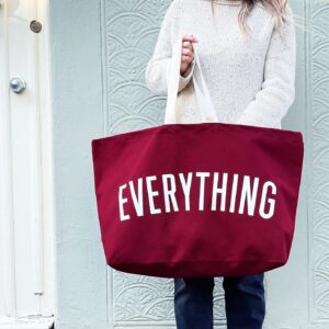 A person standing against a light-colored wall holding an extra-large burgundy canvas tote bag with the word 'EVERYTHING' printed in large white letters.