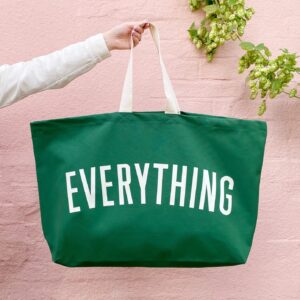 A hand holding an extra-large forest green canvas tote bag with the word 'EVERYTHING' printed in large white letters, hanging against a soft pink brick wall with greenery.