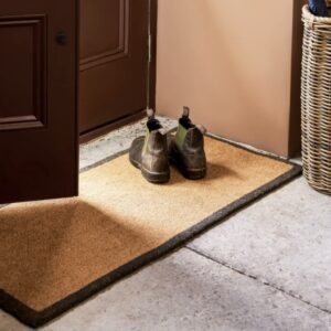 An extra-long natural coir doormat with a charcoal border, placed in front of a dark brown door, with a pair of brown boots resting on top.