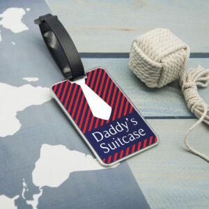 A metal luggage tag with a blue and red striped pattern and a chunky white tie design, displaying the text 'Daddy's Suitcase', attached with a black strap.