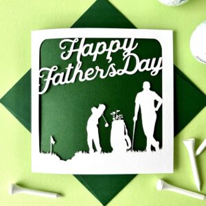 Golf Papercut Father's Day Card: A square Father’s Day card featuring a white laser-cut silhouette of a father and child playing golf against a dark green background. The card is styled on a light green surface with white golf tees and a green envelope.