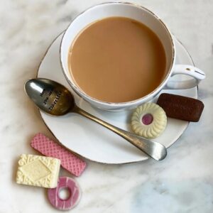 Handmade Chocolate British Biscuits: A cup of tea with milk on a white saucer, surrounded by five realistic mini chocolates shaped like classic British biscuits including a custard cream, bourbon, party ring, jammie ring, and pink wafer, next to a silver spoon engraved with "Mum you are TEA-riffic."