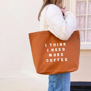 A person wearing a white quilted jacket carries a tan, 100% cotton canvas tote bag featuring the text 'I THINK I NEED MORE COFFEE' in off-white lettering.