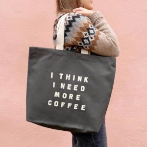 A person wearing a patterned knit sweater carries a dark grey, 100% cotton canvas tote bag featuring the text 'I THINK I NEED MORE COFFEE' in off-white lettering.