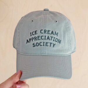Hand holding a pale vintage blue cotton baseball cap with Ice Cream Appreciation Society embroidered on the front, set against a light wooden background.