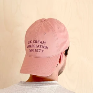 Back view of a person wearing a dusty rose cotton baseball cap with "ICE CREAM APPRECIATION SOCIETY" embroidered in mauve on the back, above an adjustable cotton strap.