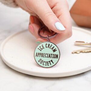 A close-up shot of a hand holding a round mint green enamel keyring with black text that reads Ice Cream Appreciation Society, featuring a matching black-toned lobster clasp and keyring.