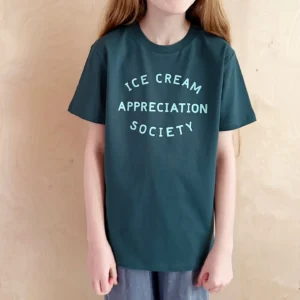 A child modeling a deep teal kid’s t-shirt featuring the text "Ice Cream Appreciation Society" printed in a soft, light mint font, set against a simple wooden background.