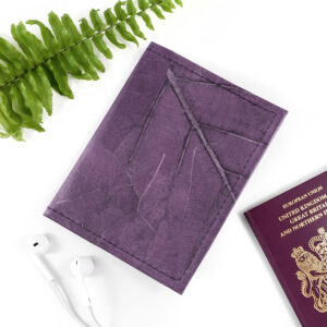 A bold purple Leaf Leather Passport Cover showcasing organic vein patterns, laid on a clean white surface next to a green fern leaf and a pair of white earphones.