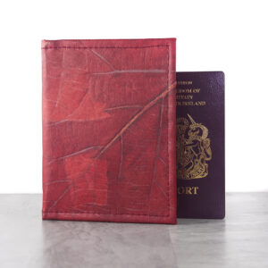 A red Leaf Leather Passport Cover made from dried, natural leaves, standing upright next to a UK passport on a light wooden surface.