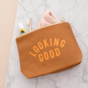 A tan canvas wash bag with the text "LOOKING GOOD" printed in bold orange letters, featuring a gold-toned zip, with a toothbrush, toothpaste, and a small pink towel tucked inside, resting on a marble surface.