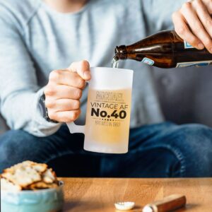 A close-up of a person holding a frosted, personalised beer tankard with the text 'MICHAEL VINTAGE AF NO.40 BREWED IN 1983' while pouring beer from a bottle into the glass at a wooden table.