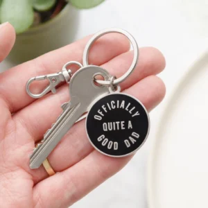 A close-up shot of a hand holding a round black enamel keyring that reads Officially Quite a Good Dad in white text, featuring a silver lobster clasp and a house key attached.
