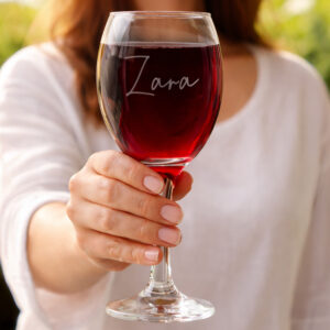 A close-up shot of a person holding a stem wine glass filled with red wine, with the name "Zara" elegantly engraved on the glass.