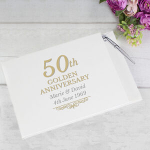 white 50th Golden Anniversary guest book with gold and grey text reading "50th GOLDEN ANNIVERSARY" with custom names "Marie & David" and date "4th June 1969", sitting on a white wooden surface next to a bouquet of purple flowers and a silver pen.
