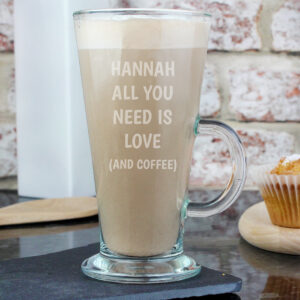 A clear glass latte filled with coffee, engraved with the name "HANNAH" and the fixed text "ALL YOU NEED IS LOVE (AND COFFEE)".