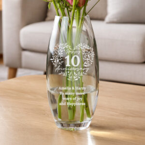 A clear, bullet-shaped glass vase sitting on a wooden table, featuring an engraved floral wreath design, the text "Happy 10 Anniversary," and names "Amelia & Harry," filled with fresh pink flowers.
